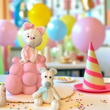 A cute pink cake topped with a teddy bear and a cat, surrounded by colorful balloons and party hats.