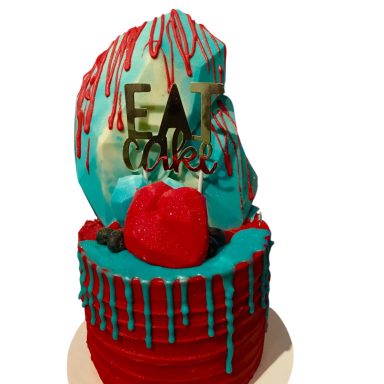 Multi-tiered cake with blue and red icing, topped with a strawberry and "EAT CAKE" sign.