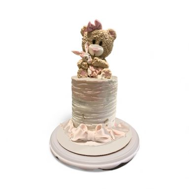 A round cake with a pink and cream design featuring a teddy bear on top.