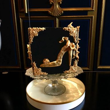 A decorative cake shaped like a high-heeled shoe, framed with ornate gold details.
