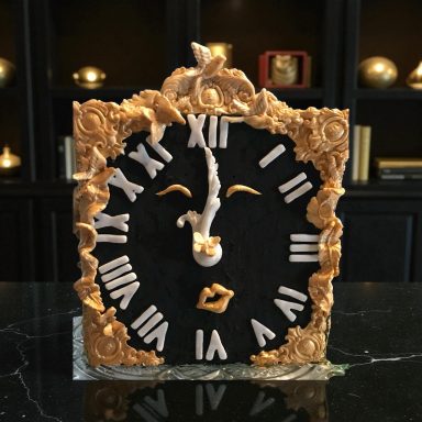 Ornate clock with Roman numerals and a face design, framed in gold and black.