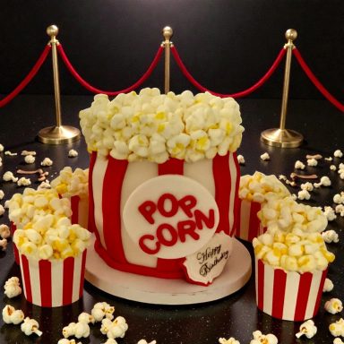 A cake designed like a popcorn bucket, surrounded by popcorn and decorative red ropes.